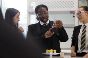 Three female students are pictured wearing safety goggles, whilst conducting a science experiment together in a lab.