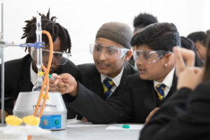 Three students are pictured wearing safety goggles, whilst conducting a science experiment together in a lab.