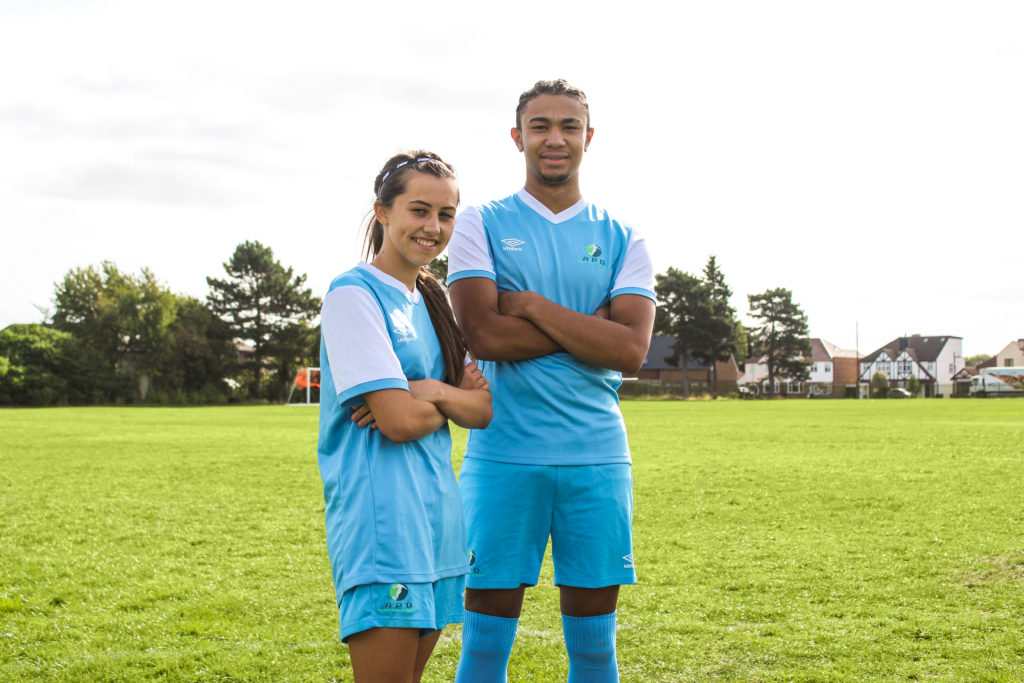 Two students are pictured standing next to each other on the school field, wearing Football kit.