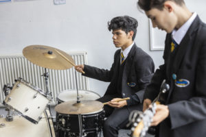 Two male students are pictured playing musical instruments alongside each other. One is playing guitar and the other is seen playing the drums.