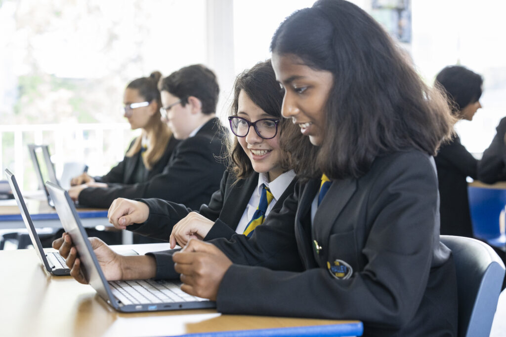 Two female Year 7 students are pictured smiling together in the foreground, as they work on laptops at their desks. Their peers can be seen working behind them in the background.