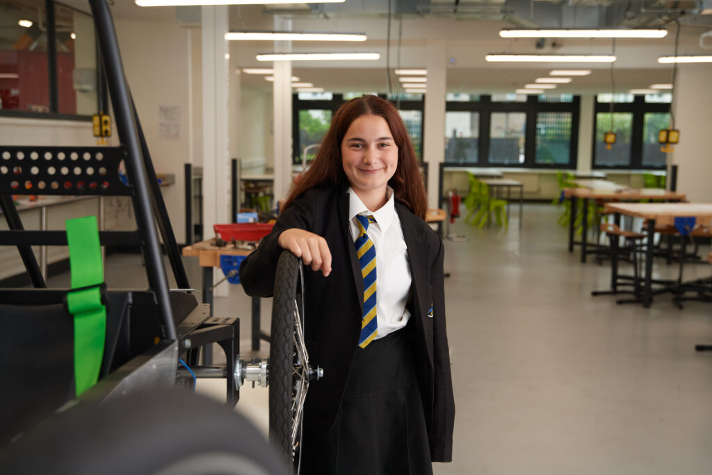 A female student can be seen smiling directly at the camera, dressed in her academy uniform, whilst standing in an open space of the academy building.