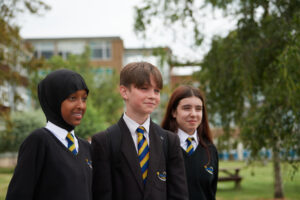 Three students, a boy and two girls, are pictured standing outdoors amongst some trees on the academy grounds, smiling for the camera.