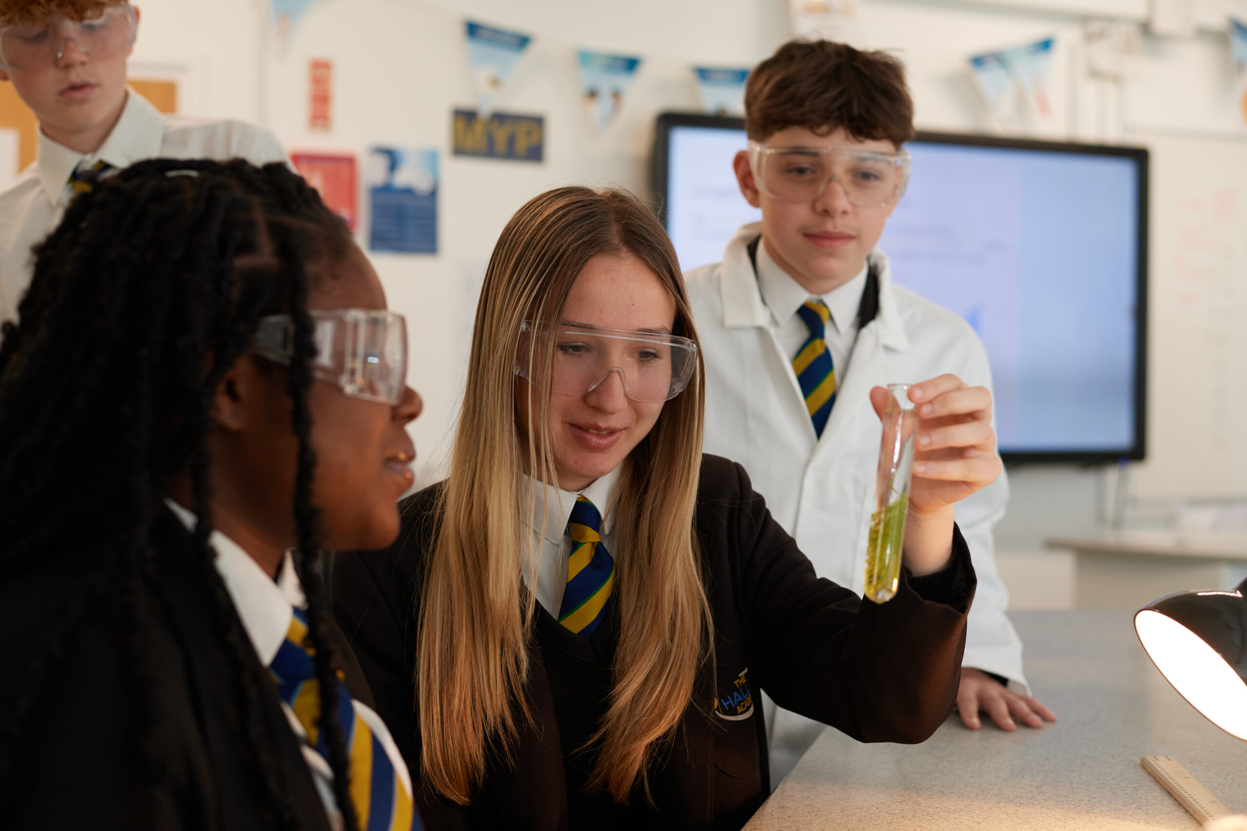 A student holding a test tube full of a yellow-green liquid, with several students sat around them, looking at the test tube